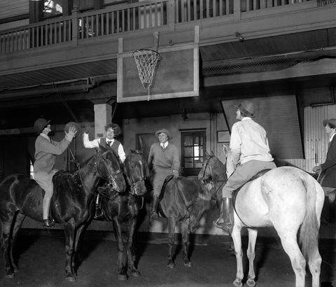 women riding horses play basketball indoors. 