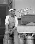 farmer sits on a milk can outside a Seed and Feed store